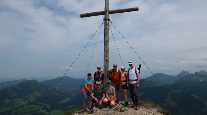 Wanderer stehen auf einem Berggipfel neben einem großen Holzkreuz mit Seilspannungen, umgeben von Berglandschaft unter bewölktem Himmel | © Christian Stadtmüller