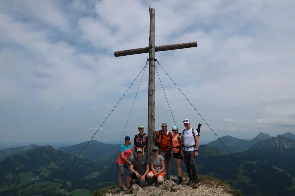 Wanderer stehen auf einem Berggipfel neben einem großen Holzkreuz mit Seilspannungen, umgeben von Berglandschaft unter bewölktem Himmel | © Christian Stadtmüller