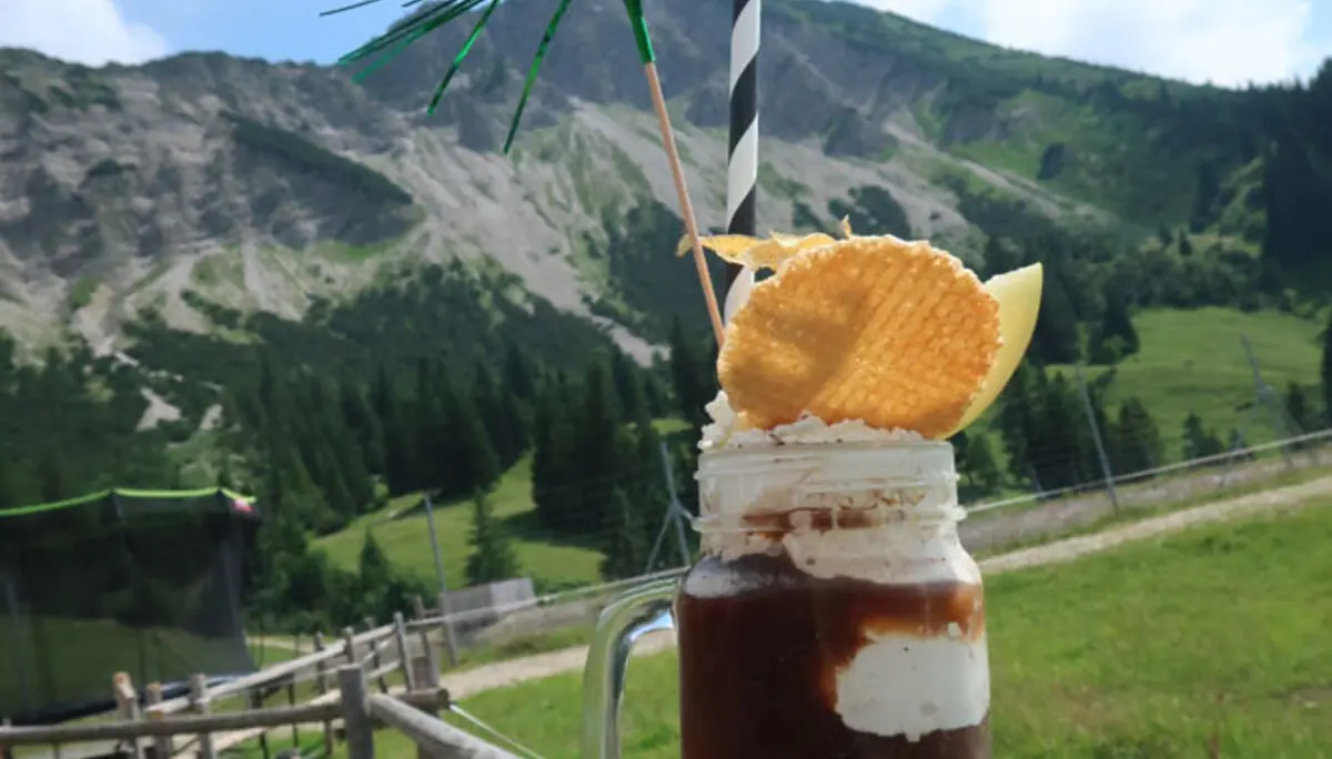 Eiskaffee mit Strohhalm auf einem Holzpfosten vor Berglandschaft | © Christian Stadtmüller