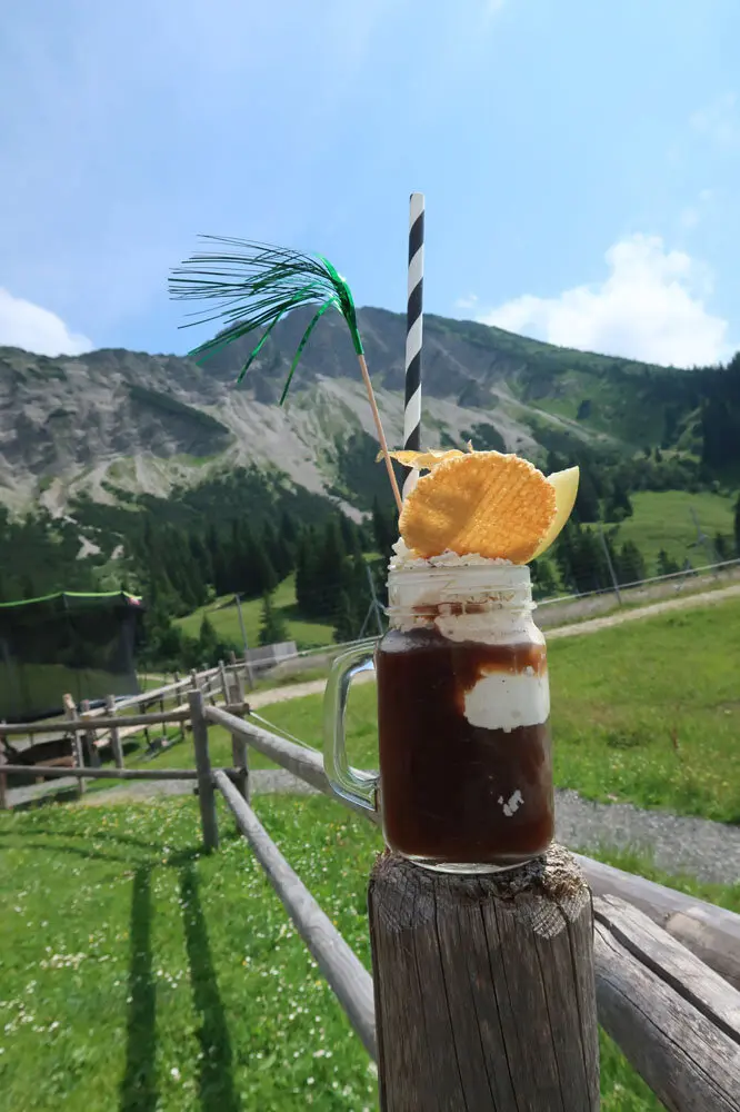 Eiskaffee mit Strohhalm auf einem Holzpfosten vor Berglandschaft | © Christian Stadtmüller
